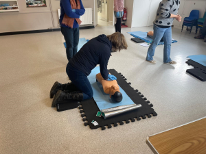 A client performing CPR on a training mannequin in a training room, supervised by a first aid instructor, demonstrating hands-on life-saving skills.